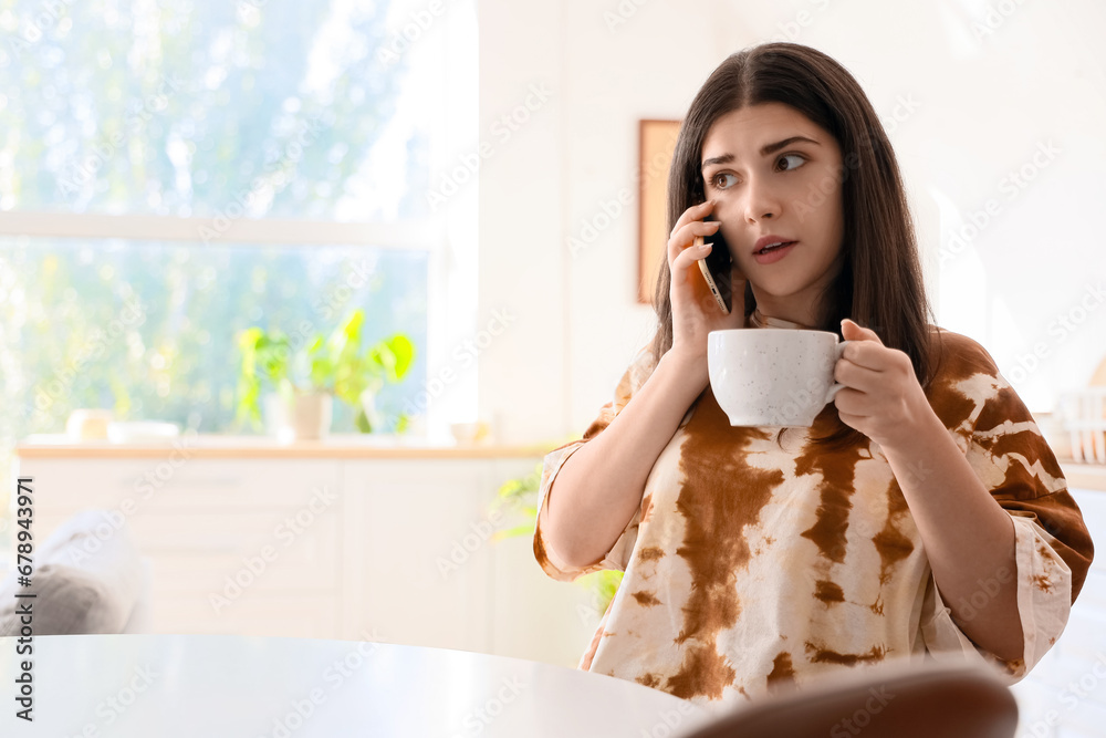 Young woman with coffee cup gossiping by mobile phone in kitchen