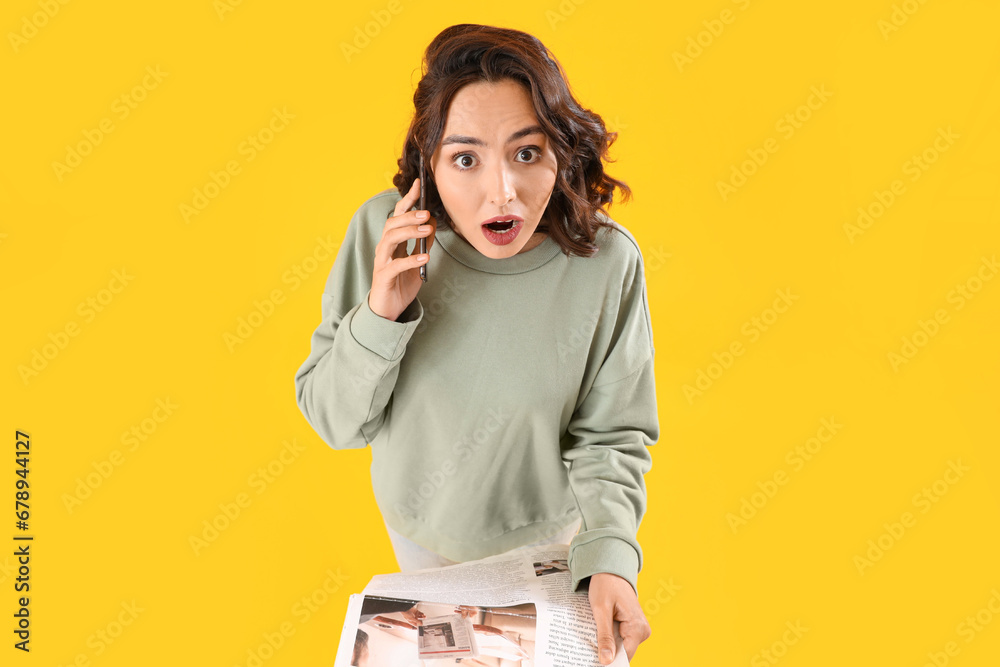 Shocked young woman with newspaper talking by mobile phone on yellow background