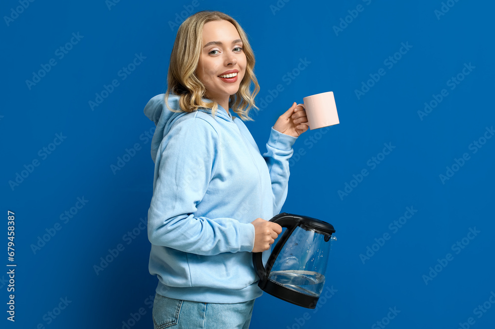 Beautiful young woman with modern electric kettle and cup on blue background