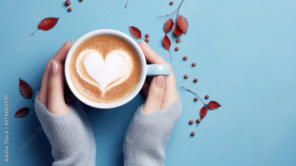 woman holding a cup of coffee, Female hands holding a cup of coffee with foam on blue background.  Hot coffee with a heart and auntumn leaves , top view