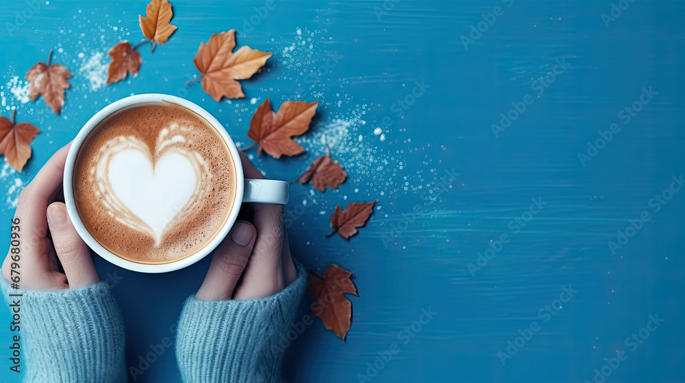 woman holding a cup of coffee, Female hands holding a cup of coffee with foam on blue background.  Hot coffee with a heart and auntumn leaves , top view