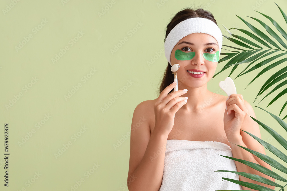 Pretty young woman with under-eye patches, guasha and face massage roller on green background with tropical leaves