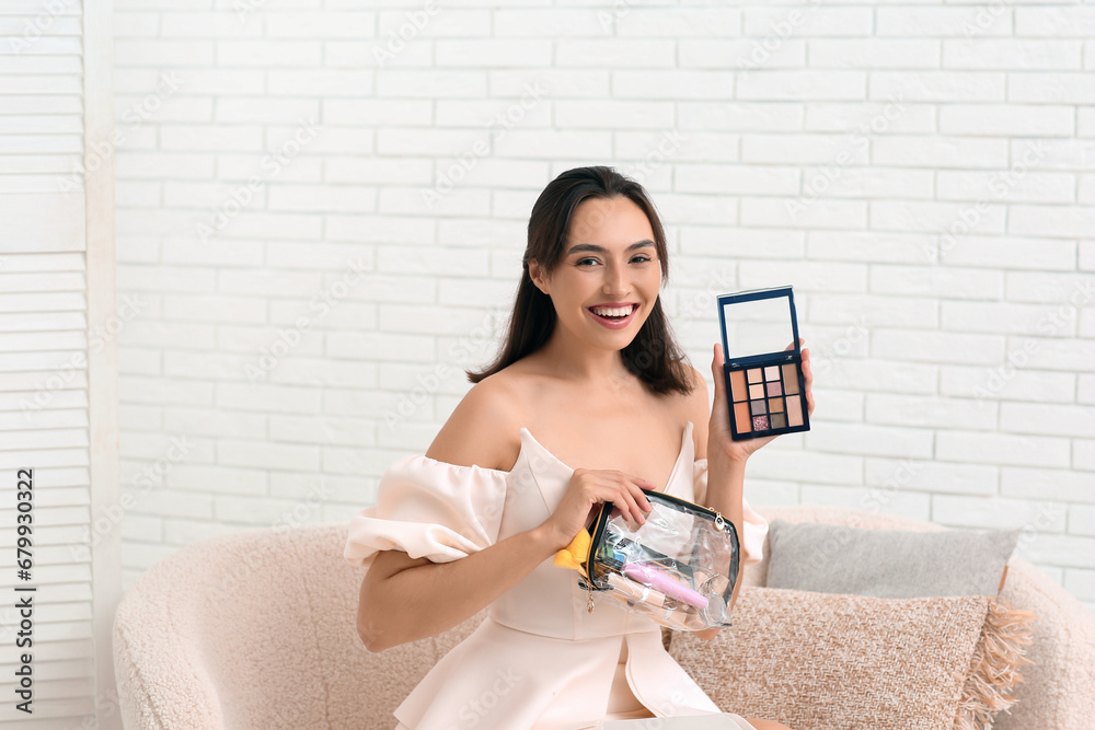 Beautiful young woman with cosmetic bag full of makeup products and eyeshadows palette in room