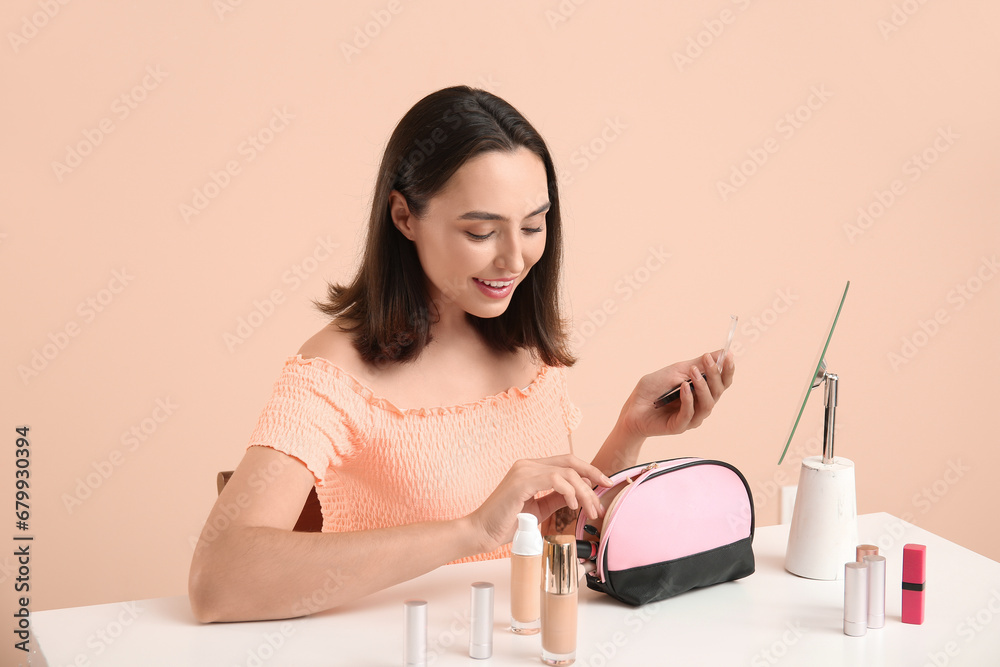 Beautiful young woman with cosmetic bag full of makeup products doing makeup at table on beige background