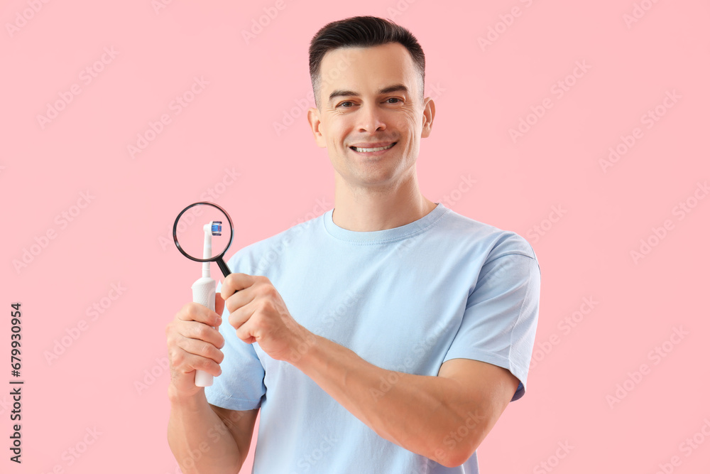 Handsome man with magnifier and electric toothbrush on pink background