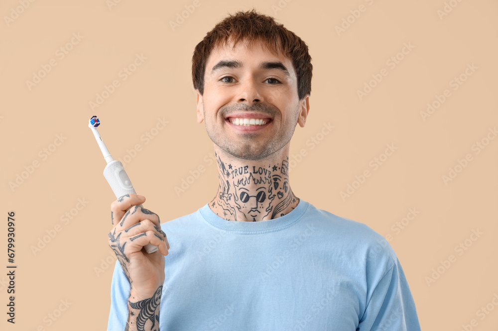 Young tattooed man with electric toothbrush on beige background