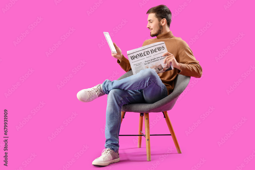 Young man with newspaper and tablet computer in armchair on purple background