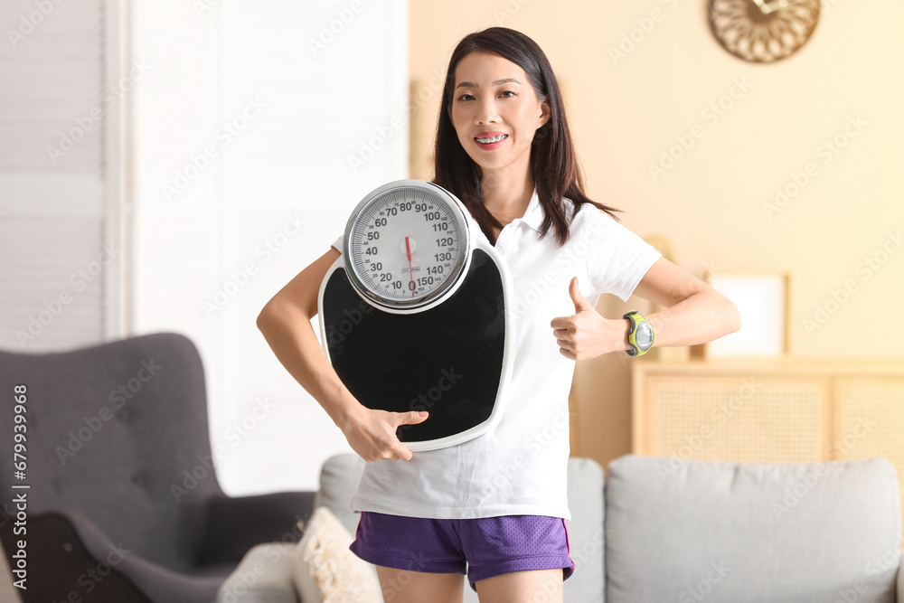 Beautiful young Asian woman with scales showing thumb-up gesture at home. Weight loss concept