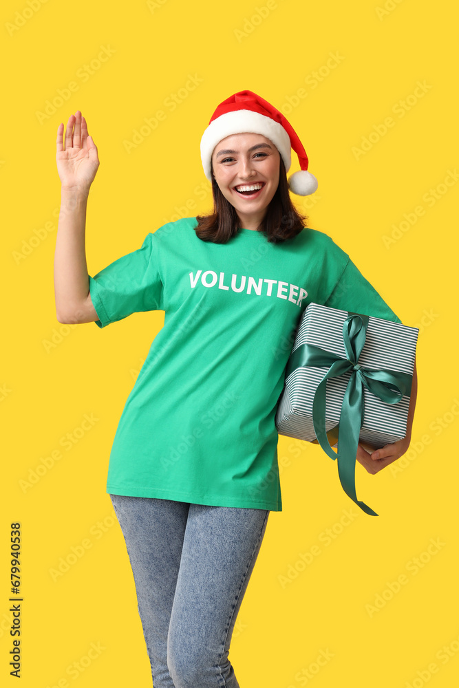 Beautiful female volunteer in Santa hat with gift box on yellow background