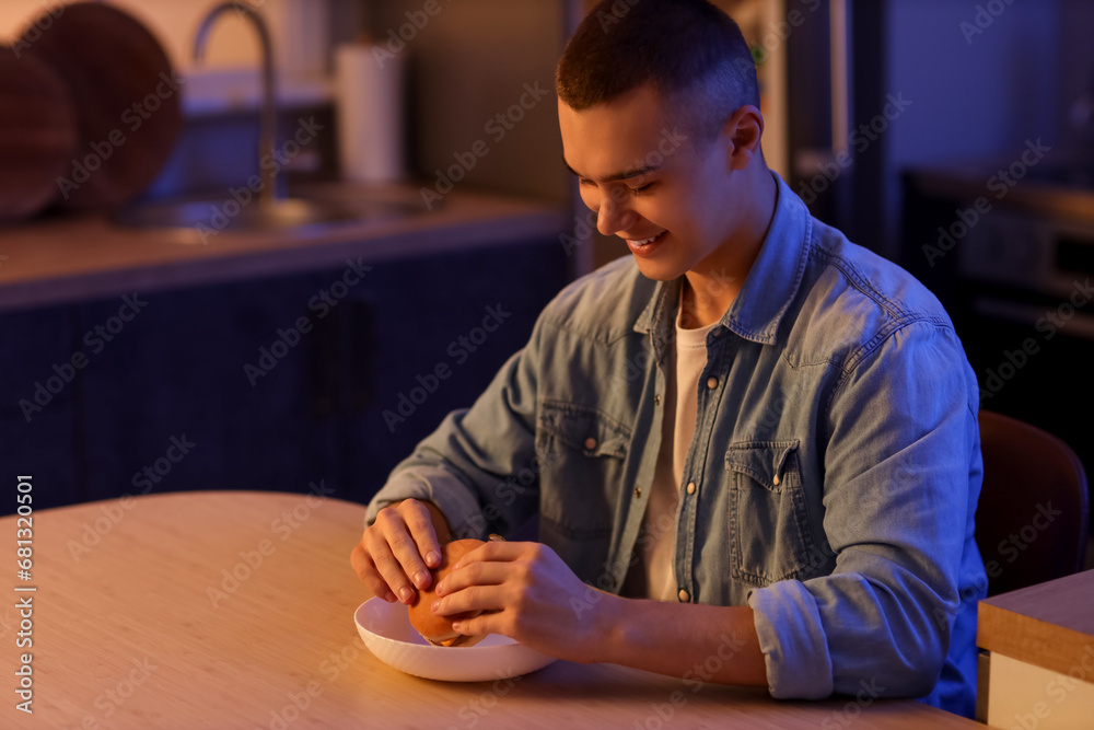 Hungry young man eating burger in kitchen at night