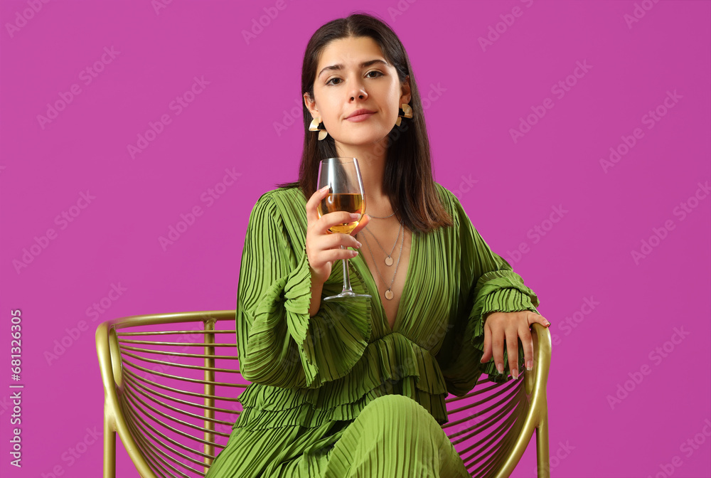 Young woman with glass of wine sitting in armchair on purple background