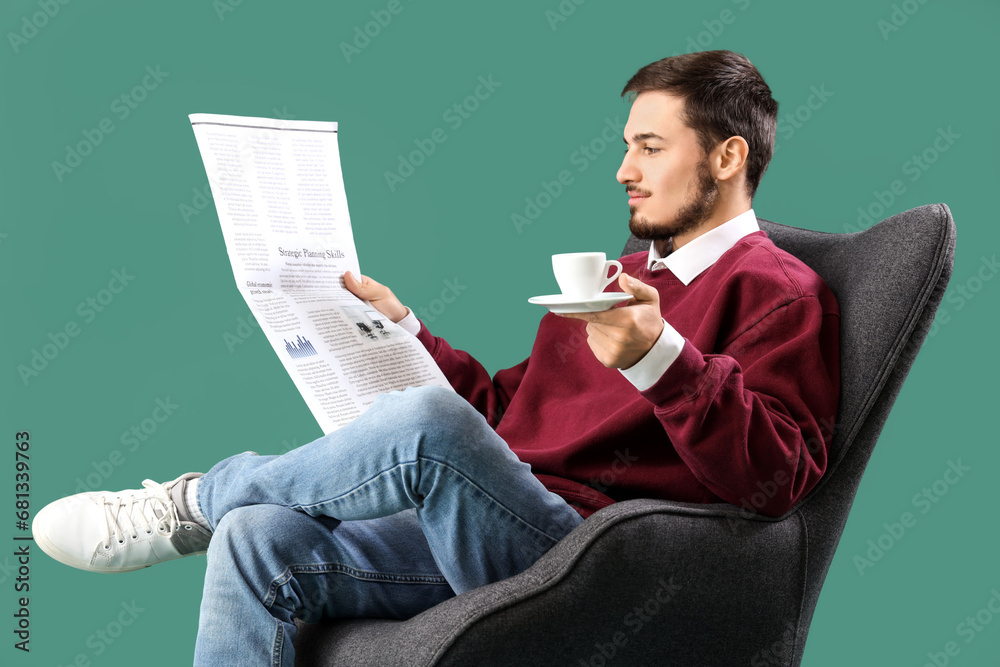 Young man with coffee cup reading newspaper in armchair on green background